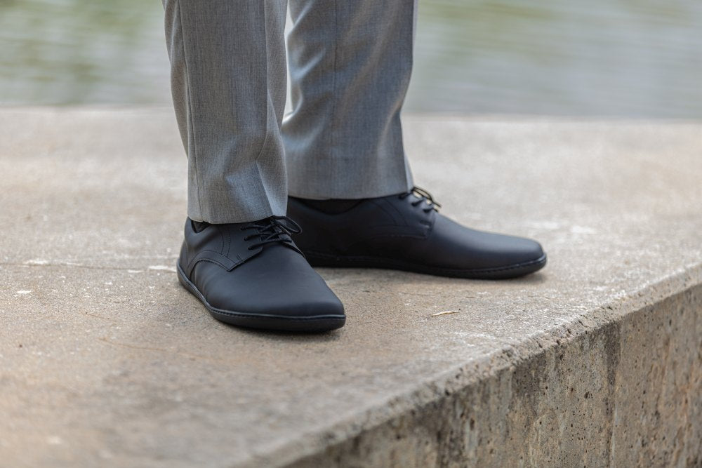Eine Person steht auf einem Betonvorsprung in der Nähe von Wasser und trägt hellgraue Anzughosen und elegante ZAQQ PEAQ Vegan Black-Schuhe. Das schwarze vegane Schuhwerk lässt auf eine bewusste Wahl in dieser lässigen, urbanen Umgebung schließen.
