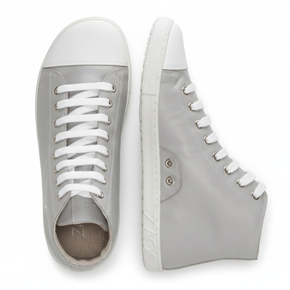 Ein Paar stylische CHUQQS Grey High-Top-Sneaker von ZAQQ mit Obermaterial aus hellgrauem Nappaleder und weißen Gummisohlen. Ein Schuh ist von der Seite zu sehen und zeigt das ZAQQ-Logo in der Nähe der Sohle, während der andere Schuh von oben zu sehen ist und seine weißen Schnürsenkel betont.