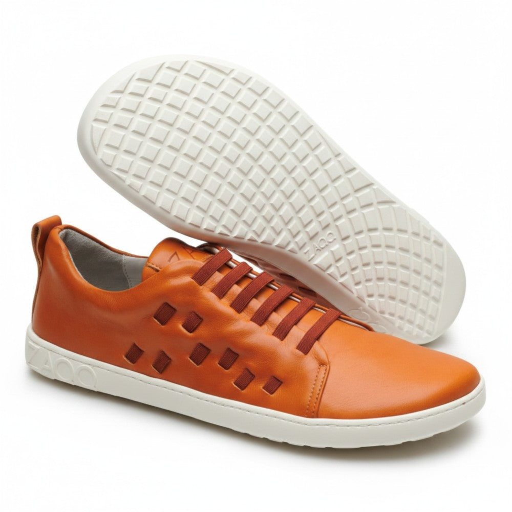 Der STRAQ Sierra von ZAQQ ist ein orangefarbener Freizeitschuh mit quadratischen Aussparungen an der Seite und einer weißen, karierten Sohle aus Nappaleder. Er fühlt sich an wie ein Barfuß-Sneaker und zeigt stolz den Markennamen sowohl auf der Ferse als auch auf der Sohle.