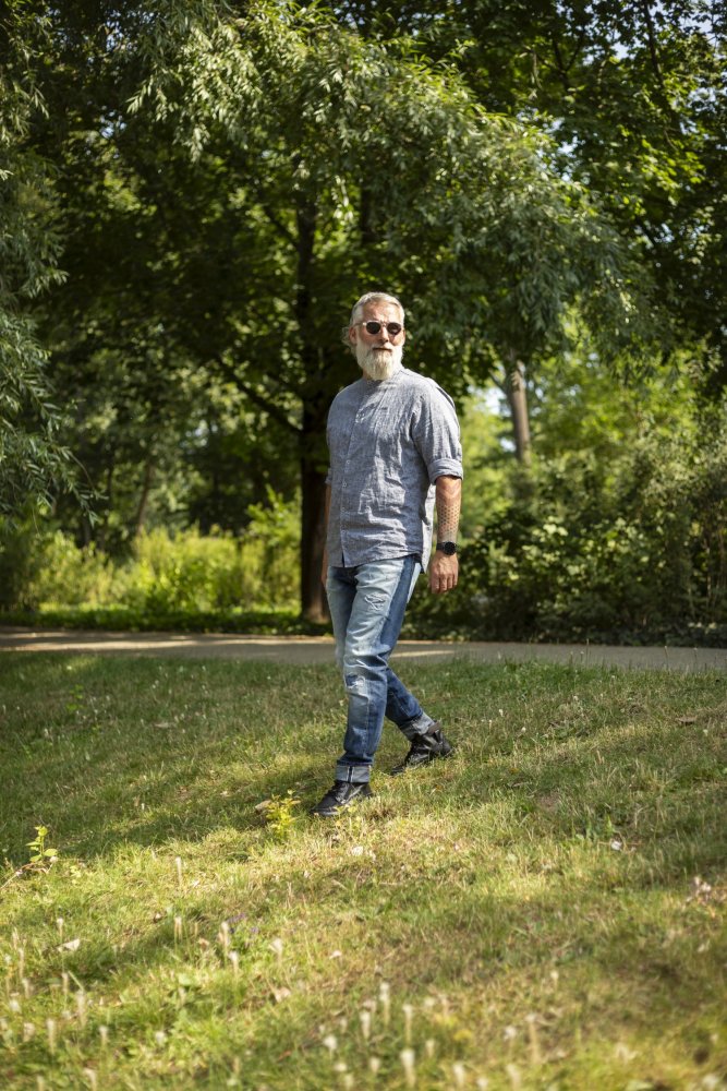 Ein Mann mit Bart und Sonnenbrille schlendert durch eine Rasenfläche im Park. Er trägt ein graues Hemd, blaue Jeans und schicke ROQA Black-Sneaker von ZAQQ. Er geht unter dem sonnigen Himmel zwischen üppigen grünen Bäumen und Sträuchern umher.